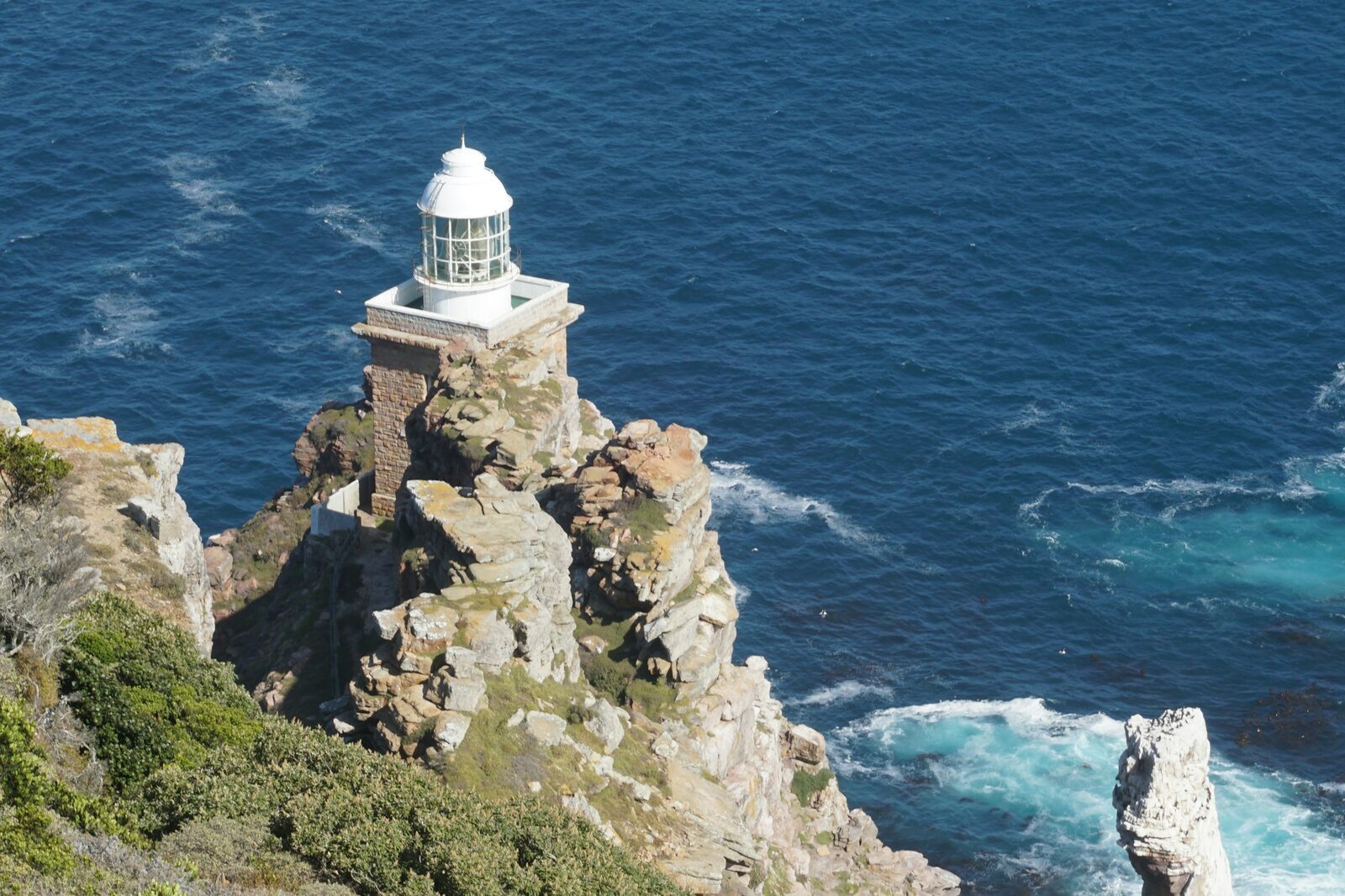 A white lighthouse on the rocky Cape coastline under a bright sky.